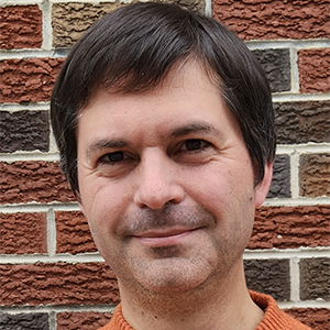 James Rising smiling in front of a brick wall while wearing an orange shirt.