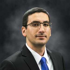 Amin Mirkouei wearing glasses, a black suit, white collared shirt, and blue tie, stands and smiles.