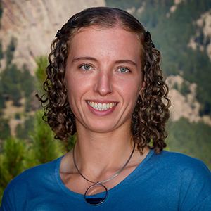 Constance Crozier stands  in front of a mountain with trees while wearing a blue shirt and smiling.